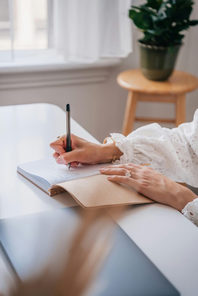 Close-up of a woman writing in a notebook at a bright home office desk.